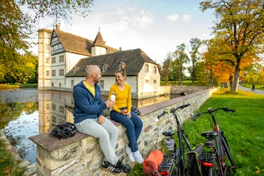 Willebadessen-Wasserschloss Schweckhausen-Teutoburger-Wald-Tourismus-D-Ketz-106.jpg Ein Paar sitzt auf einer Mauer am Wasser, genießt Eis vor einem historischen Gebäude mit Fahrrädern.