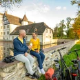 Willebadessen-Wasserschloss Schweckhausen-Teutoburger-Wald-Tourismus-D-Ketz-106.jpg Ein Paar sitzt auf einer Mauer am Wasser, genießt Eis vor einem historischen Gebäude mit Fahrrädern.