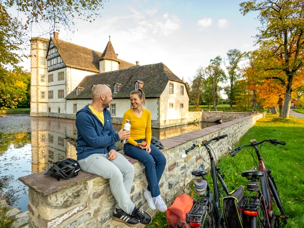 Willebadessen-Wasserschloss Schweckhausen-Teutoburger-Wald-Tourismus-D-Ketz-106.jpg Ein Paar sitzt auf einer Mauer am Wasser, genießt Eis vor einem historischen Gebäude mit Fahrrädern.