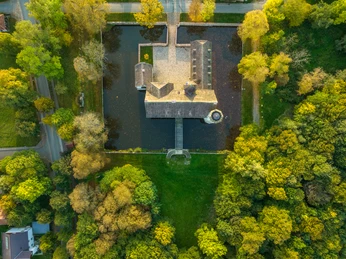 Luftaufnahme einer von Bäumen umgebenen historischen Wasserburg mit rechteckigem Graben und Zufahrtsbrücke.