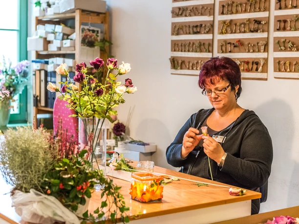 Duitse kunstbloem Sebnitz Een vrouw zit aan een tafel en schikt bloemen; om haar heen staan vazen met kleurrijke bloemen en planten. De sfeer is creatief en gefocust.