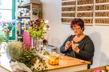 German artificial flower Sebnitz A woman sits at a table and arranges flowers; around her are vases with colorful flowers and plants. The atmosphere is creative and concentrated.