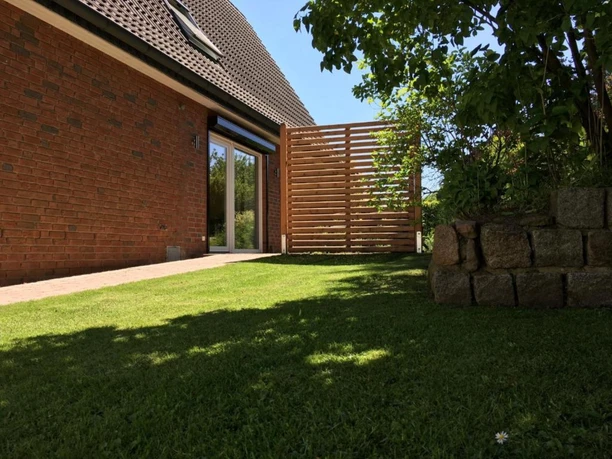 Außenansicht Backsteingebäude mit Terrassentür, gepflegtem Rasen, Holzsichtschutz und kleiner Steinmauer.