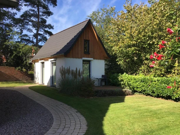Ferienhaus Kleines Haus mit grauem Dach in grünem Garten, umgeben von Bäumen und Blumen unter blauem Himmel.
