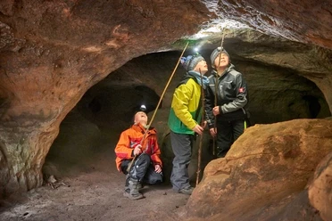Cave Tour Quirl Three people with headlamps and hiking poles explore a cave, looking curiously at the ceiling; they are wearing colorful outdoor clothing.