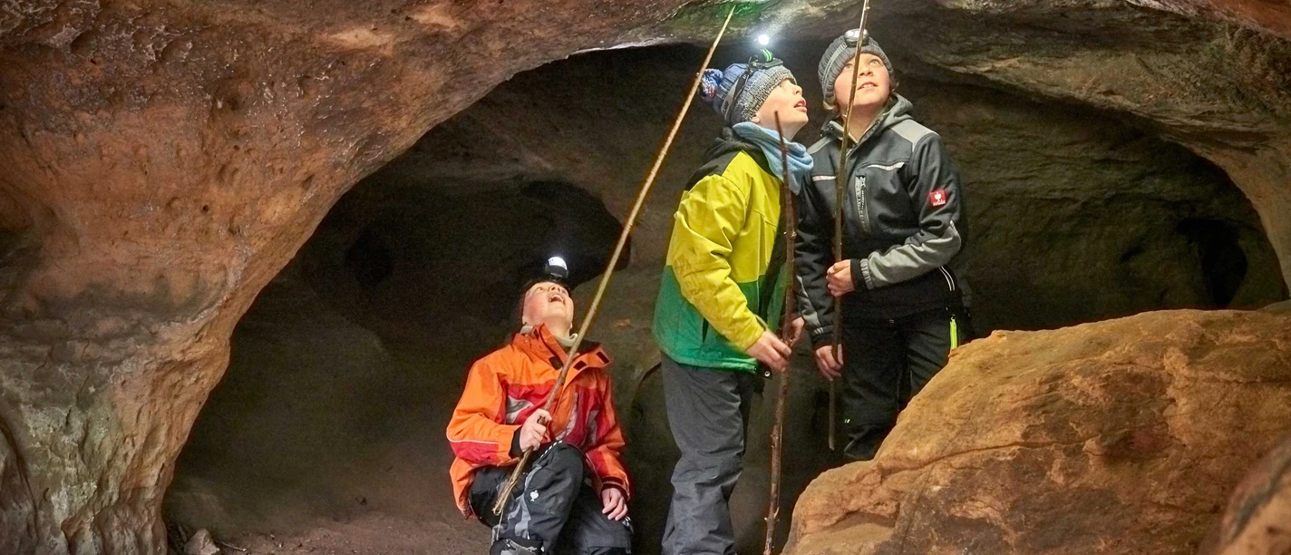 Höhlentour Quirl Drei Personen mit Stirnlampen und Wanderstöcken erkunden eine Höhle, schauen neugierig zur Decke; sie tragen bunte Outdoor-Kleidung.