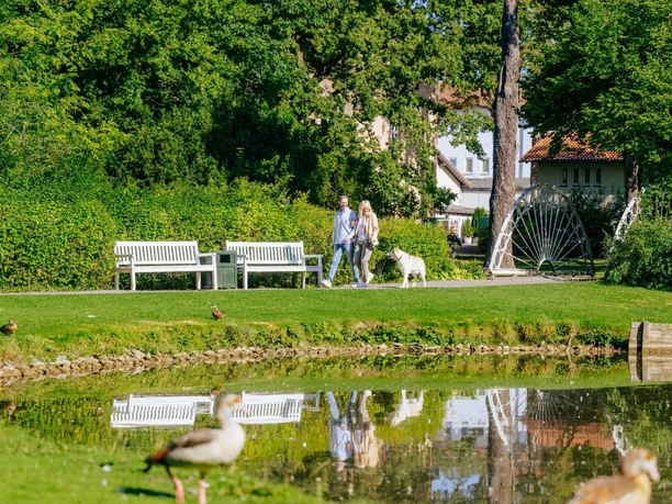 Der Ententeich im Gräflichen Park wo ein Paar im Hintergrung mit ihrem Hund entlang gehen.