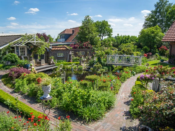 Garten_Privatgarten_Mueller Bollenhagen-32.jpg Ein idyllischer Garten mit blühenden Blumen, einem Teich und einer weißen Brücke.
