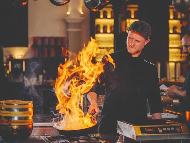 Mann brät im Restaurant Pferdestall das Essen, Feuer kommt aus der Pfanne raus.
