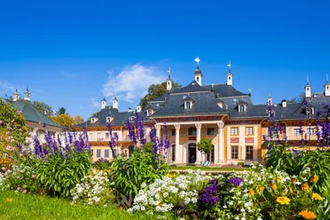 Pillnitz Castle Pillnitz Palace with its yellow façade and dark roof, surrounded by colorful flowerbeds under a clear blue sky.