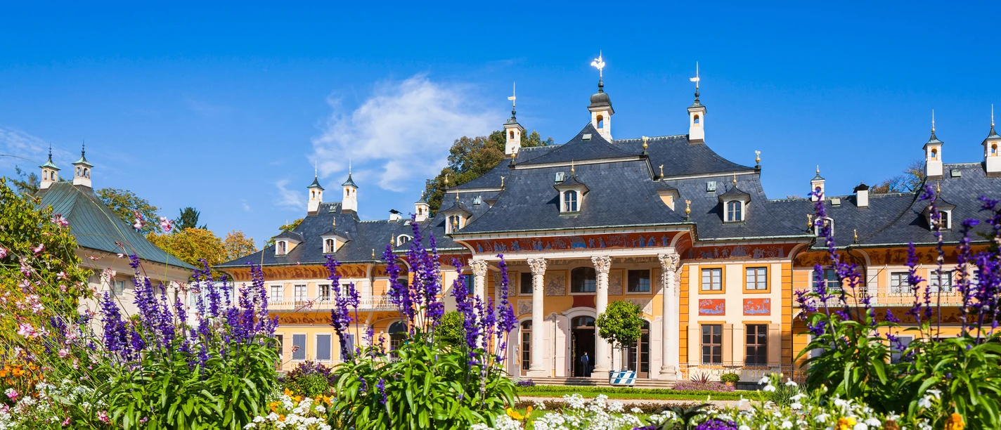 Schloss Pillnitz Schloss Pillnitz mit gelber Fassade und dunklem Dach, umgeben von bunten Blumenbeeten unter klarem, blauem Himmel.