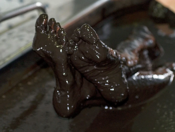 Moorfüße in der Wanne Füße, die vollständig mit dunklem Moor bedeckt sind, ragen aus einer Wanne mit brauner Moorflüssigkeit.