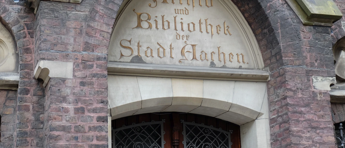 Grashaus Tür in einem steinernen Torbogen, in den in alter Schrift "Archiv und Bibliothek der Stadt Aachen" eingemeißelt ist.