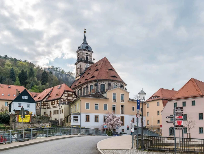Stadtkirche Königstein Straßenszene mit Fachwerkhäusern und einer Kirche mit Turm, umgeben von Hügeln und bewölktem Himmel; frühlingshafte Atmosphäre.