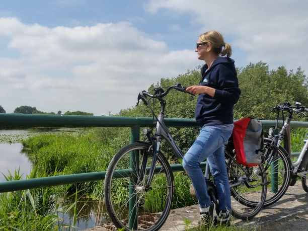 Radfahren in der Hammeniederung A cyclist stands next to her bike on a bridge and looks out over a landscape with a lake.