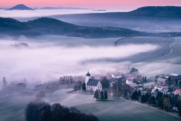 Sonnenaufgang auf dem Papststein mit Blick nach Papstdorf Misty valley with a white church and surrounding houses, surrounded by fields and hills in the morning light, under a pink sky.