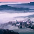 Sonnenaufgang auf dem Papststein mit Blick nach Papstdorf Nebliges Tal mit einer weißen Kirche und umliegenden Häusern, umgeben von Feldern und Hügeln im Morgenlicht, unter einem rosa Himmel.