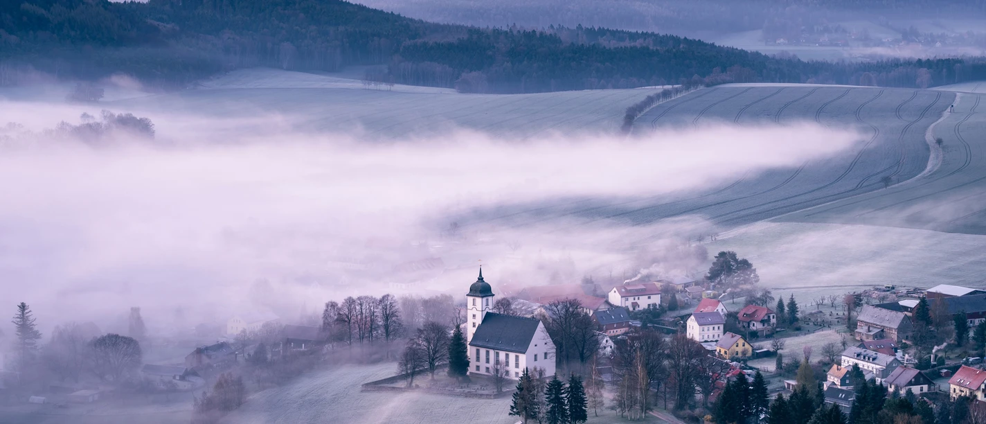 Sonnenaufgang auf dem Papststein mit Blick nach Papstdorf Misty valley with a white church and surrounding houses, surrounded by fields and hills in the morning light, under a pink sky.