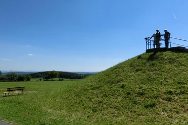 Oelsener Höhe viewing platform Two people are standing on a viewing platform on a grassy hill, surrounded by a vast, green landscape under a blue sky.