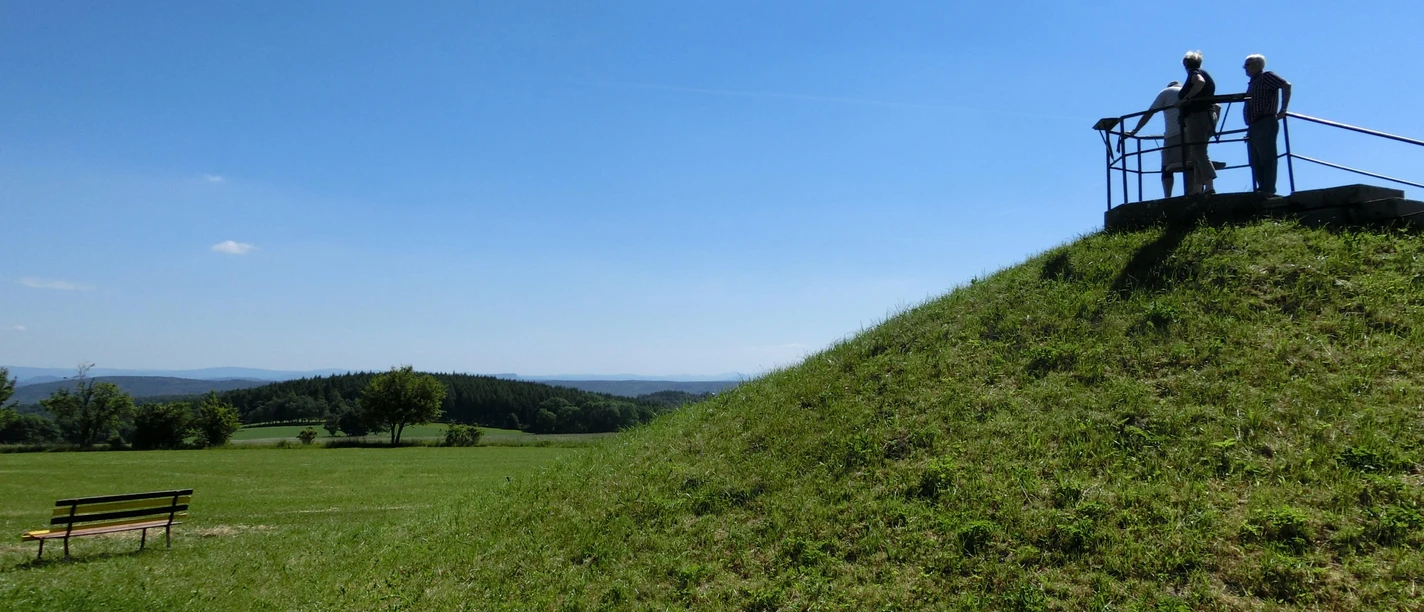 Oelsener Höhe viewing platform Two people are standing on a viewing platform on a grassy hill, surrounded by a vast, green landscape under a blue sky.