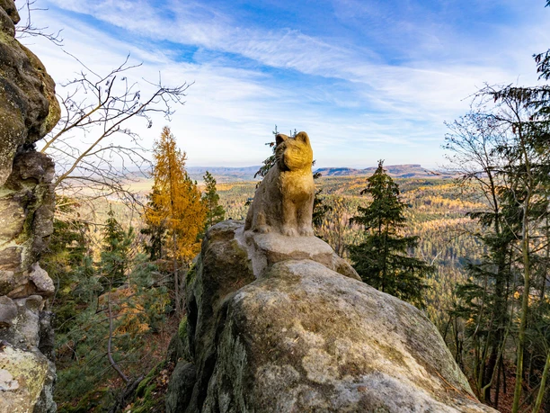 Katzfels Steinerne Luchsstatue auf einem Felsen mit Blick auf einen herbstlichen Wald und blauen Himmel im Hintergrund.