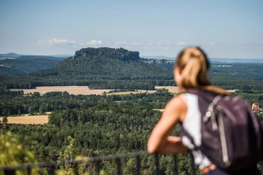 Brandaussicht Een vrouw met rugzak kijkt uit over een groen, heuvelachtig landschap met een opvallende rots op de achtergrond onder een heldere hemel.