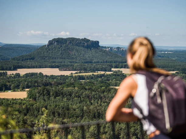Brandaussicht A woman with a rucksack looks out over a green, hilly landscape with a striking rock in the background under a clear sky.