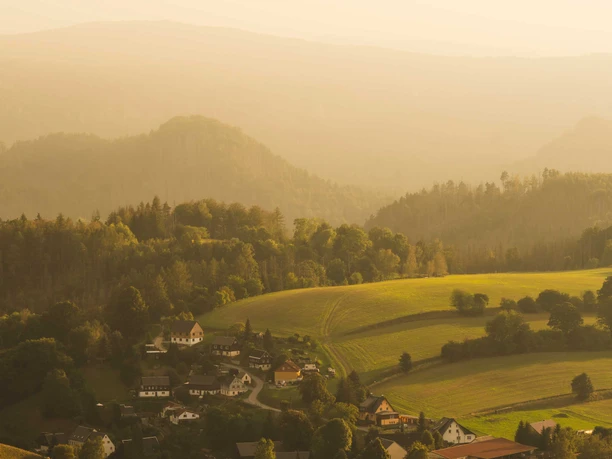 Pohled z Wachbergu Malá vesnička zasazená do zelených kopců a lesů, ozářená teplým zlatavým sluncem v tiché a klidné atmosféře.