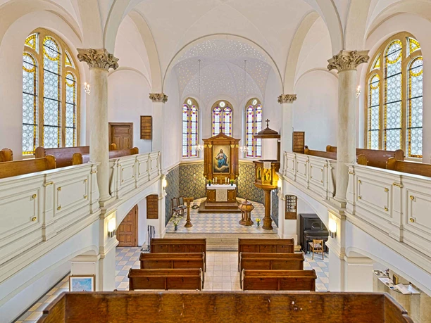 Binnenzicht van een kerk met witte muren, kleurrijke glas-in-loodramen, houten banken en een altaar met een schilderij. Heldere, rustige sfeer.