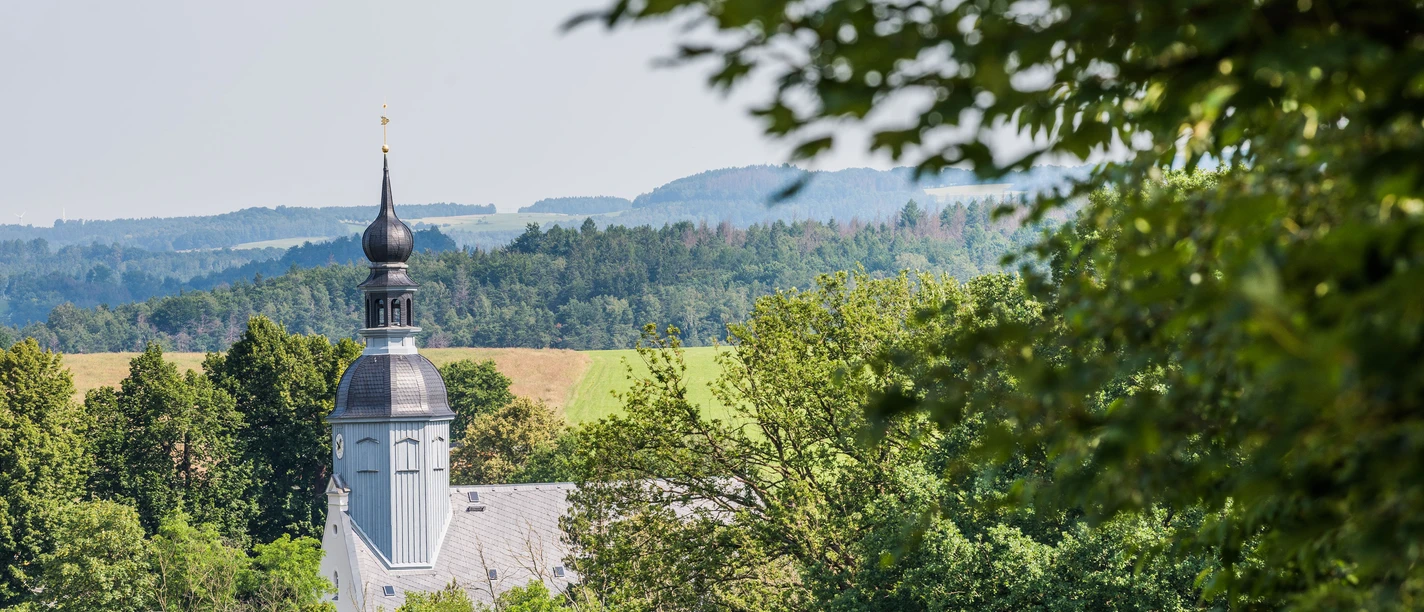 Kirche Reinhardtsdorf A church with a black onion dome rises out of the dense greenery, with rolling hills under a clear sky in the background.