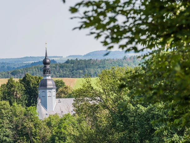 Kirche Reinhardtsdorf Eine Kirche mit einem schwarzen Zwiebelturm ragt aus dichtem Grün hervor, im Hintergrund sanfte Hügel unter klarem Himmel.