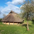 Das Eisenzeithaus zwischen blühenden Obstbäumen.jpg Nachgebautes steinzeitliches Langhaus auf grüner Wiese, umgeben von Bäumen.