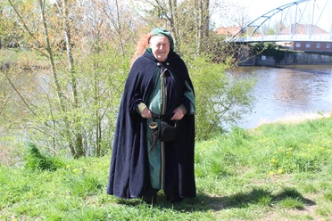 Stadtführer Nienburg Eine mittelalterlich gekleidete Person steht vor einem Fluss, hinter einer modernen Brücke.