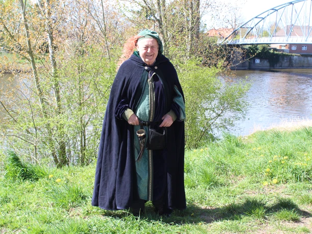 Stadtführer Nienburg Eine mittelalterlich gekleidete Person steht vor einem Fluss, hinter einer modernen Brücke.