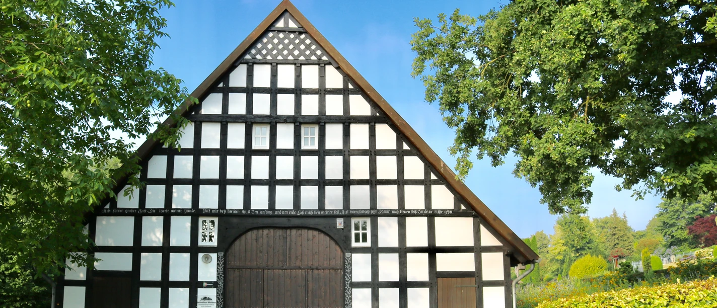 Historisches Fachwerkhaus mit symmetrischem Design, umgeben von Bäumen und gepflegtem Rasen.