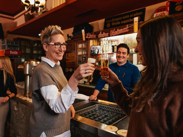 Früh im Veedel Scene in a rustic brewery: two women toast with Kölsch beer.