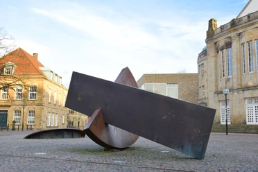 “Gleiches Gewicht – Gleichgewicht” von Joachim Bandau Skulptur aus rostigem Metall auf einem gepflasterten Platz in Osnabrück umgeben von Gebäuden.