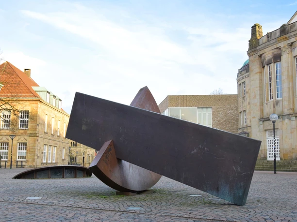 “Gleiches Gewicht – Gleichgewicht” von Joachim Bandau Skulptur aus rostigem Metall auf einem gepflasterten Platz in Osnabrück umgeben von Gebäuden.