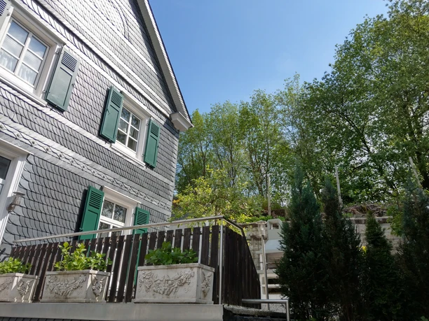 IMAG0375.jpg Historisches Haus mit grünen Fensterläden und Schieferfassade, umgeben von hohen Bäumen.