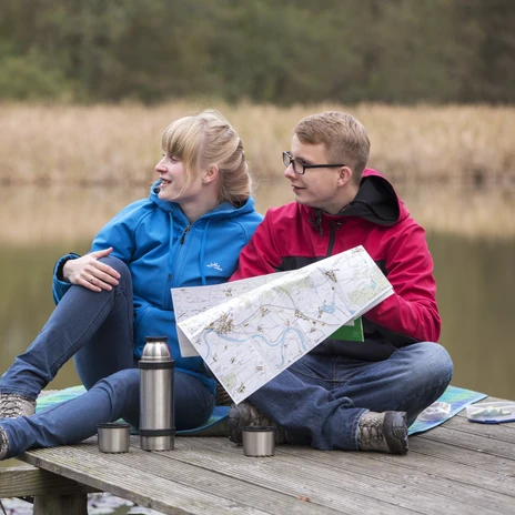 Ein Paar sitzt am Steg eines Gewässers in der Dahlener Heide mit einer Karte in der Hand, im Hintergrund ein GetreidefeldA couple sits on the jetty of a body of water in the Dahlener Heide with a map in their hands, a field of grain in the background.Pár sedí na molu u vodní plochy v Dahlen Heath s mapou v ruce, v pozadí je pole s obilím.Para siedzi na nabrzeżu zbiornika wodnego w Dahlen Heath z mapą w rękach, w tle pole zbożaEen echtpaar zit op de steiger van een waterlichaam in de Dahlen Heide met een kaart in hun handen, een graanveld op de achtergrondUna coppia seduta sul pontile di uno specchio d'acqua nella brughiera di Dahlen con una mappa in mano, un campo di grano sullo sfondo
