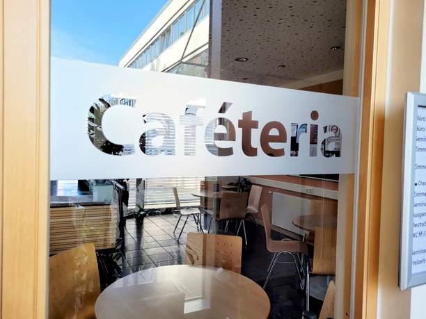 Transparente Glastür mit Schriftzug „Caféteria“, Holzmöbel und runde Tischanordnung innen.