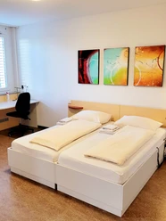 Doppelzimmer mit zwei Einzelbetten, einem Schreibtischstuhl und drei abstrakten Wandbildern.