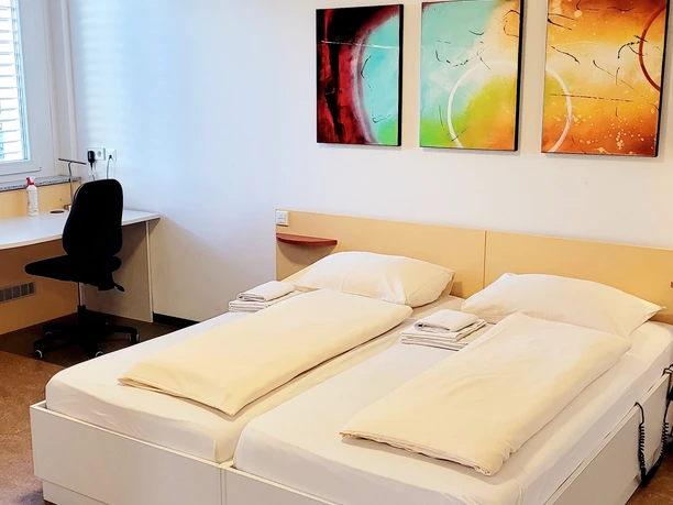 Doppelzimmer mit zwei Einzelbetten, einem Schreibtischstuhl und drei abstrakten Wandbildern.
