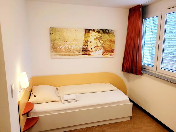 Ein helles Einzelzimmer mit einem Bett, Wandbild eines Schiffs, und einem Fenster mit orangen Vorhängen.