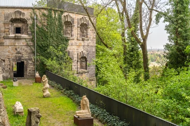 Alte Steinmauer des Schloss Sonnenstein mit Efeu bewachsen, umgeben von Bäumen; im Vordergrund stehen mehrere antike Skulpturen auf einer grünen Wiese.