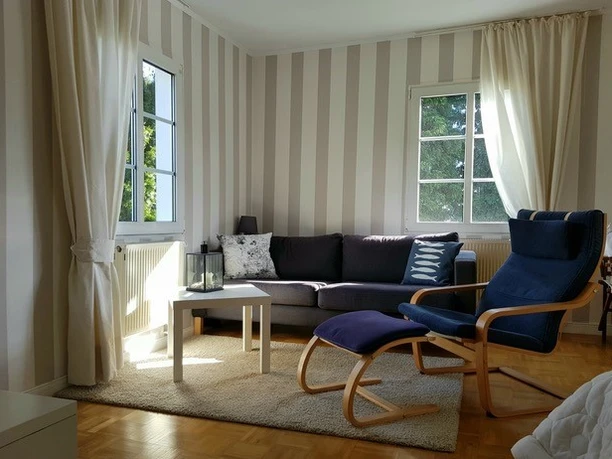 Wohnzimmer.jpg Helles Wohnzimmer mit gemütlicher Sitzgruppe, gestreiften Tapeten, großem Fenster und Holzboden.