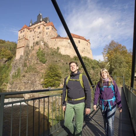 A man and a woman in hiking clothes walk across a bridge over the Zschopau, with Kriebstein Castle in the background surrounded by woods