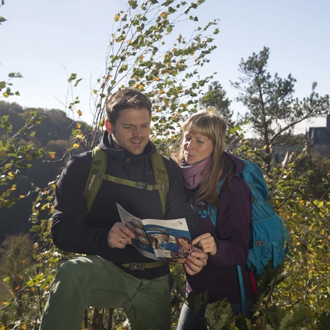A man and a woman in hiking gear look at a brochure together in the countryside, Kriebstein Castle in the background