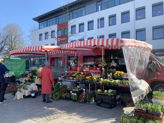 wochenmarkt-bad-zwischenahn-stand.jpg Ein Marktstand auf dem Wochenmarkt Bad Zwischenahn bietet unter einem roten, gestreiften Vordach eine Vielfalt an bunten Blumen und Pflanzen an. Im Vordergrund ist eine Kundin in rotem Mantel zu sehen, die Ware begutachtet, während ein weiteres Zelt im Hintergrund steht.