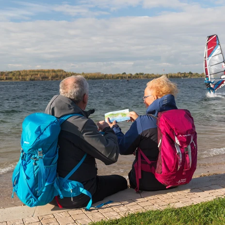 Rear view of an elderly couple looking at a photo on the edge of Lake Schladitz, surfers, boats, forests, blue sky
